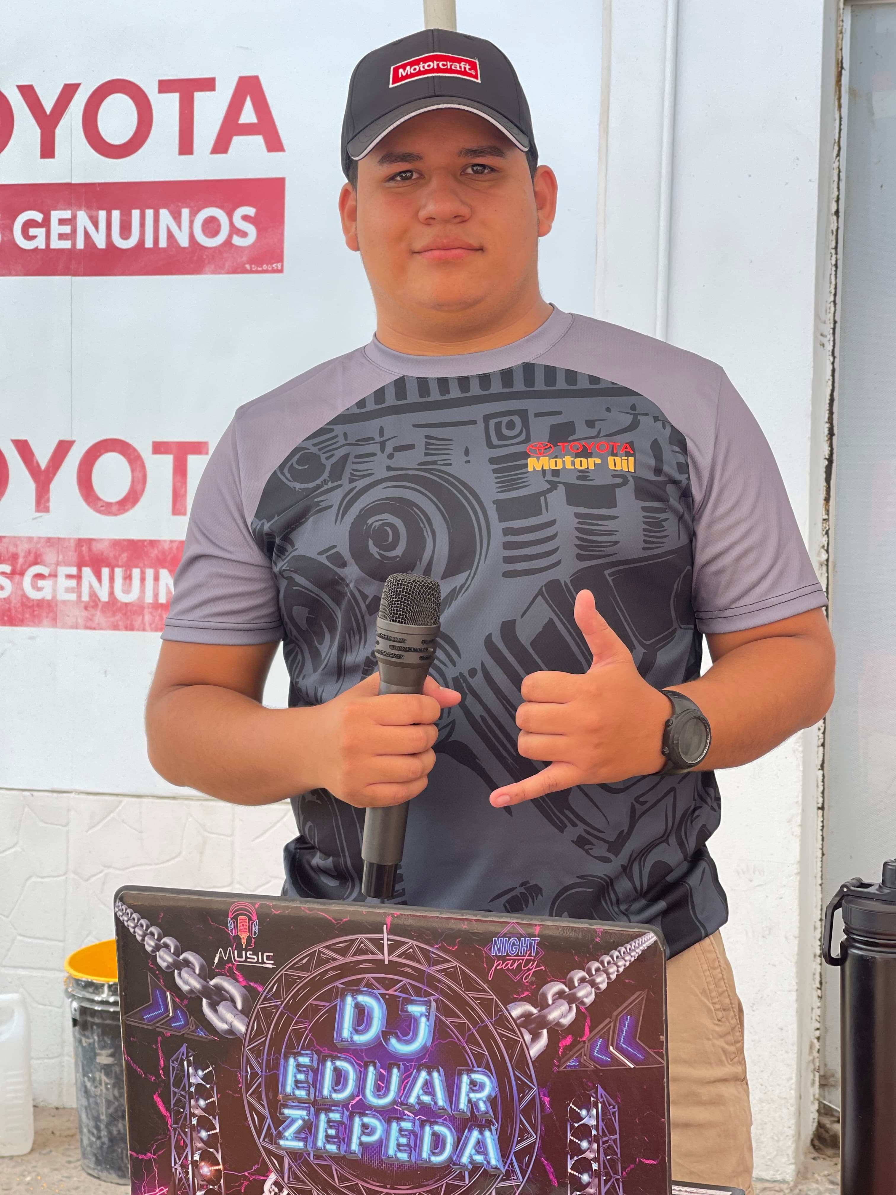 DJ Eduar Zepeda con micrófono y gesto de mano frente a su laptop personalizada.