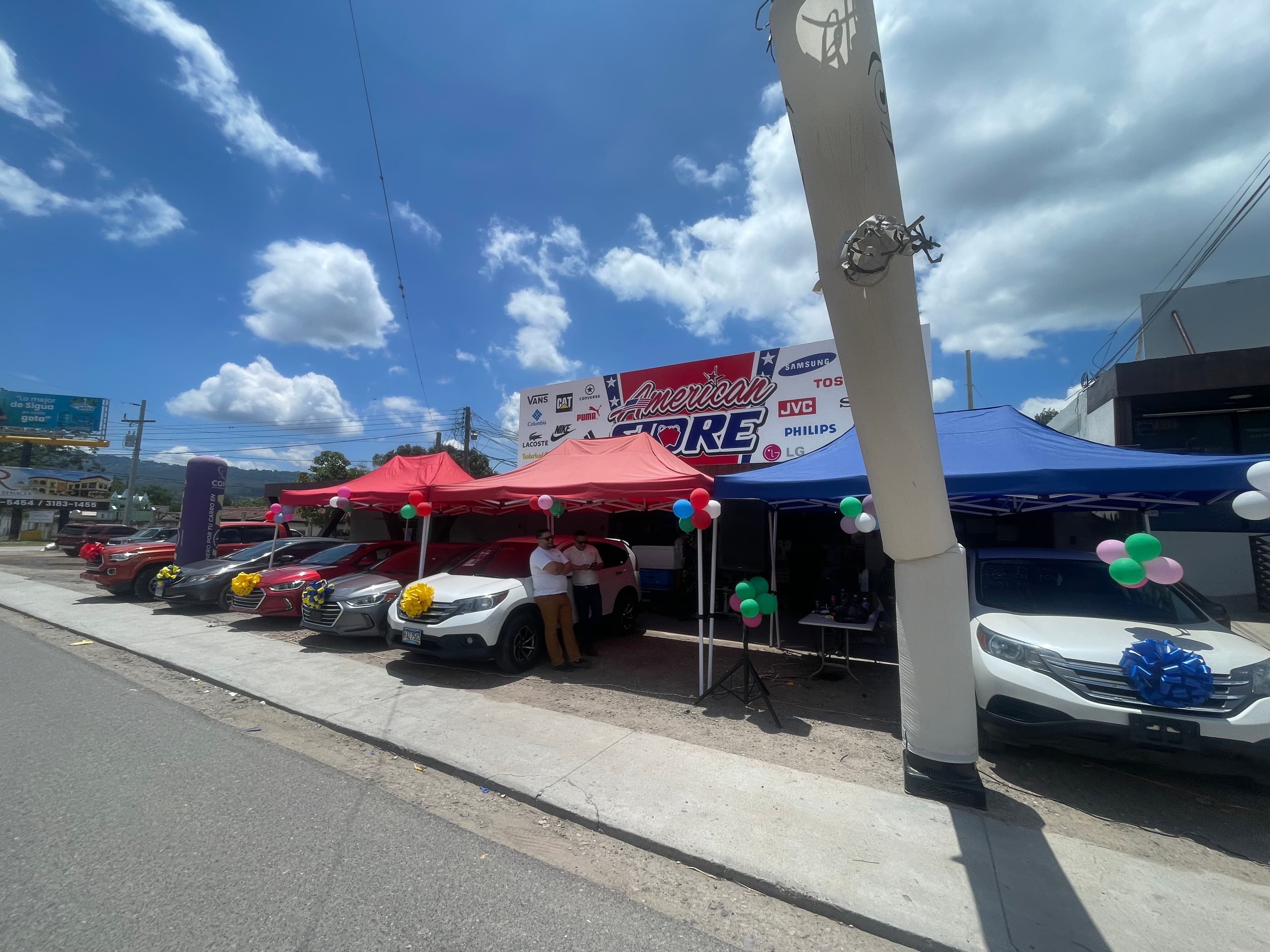 Autos en venta bajo carpas coloridas con globos y lazos frente a American Store.