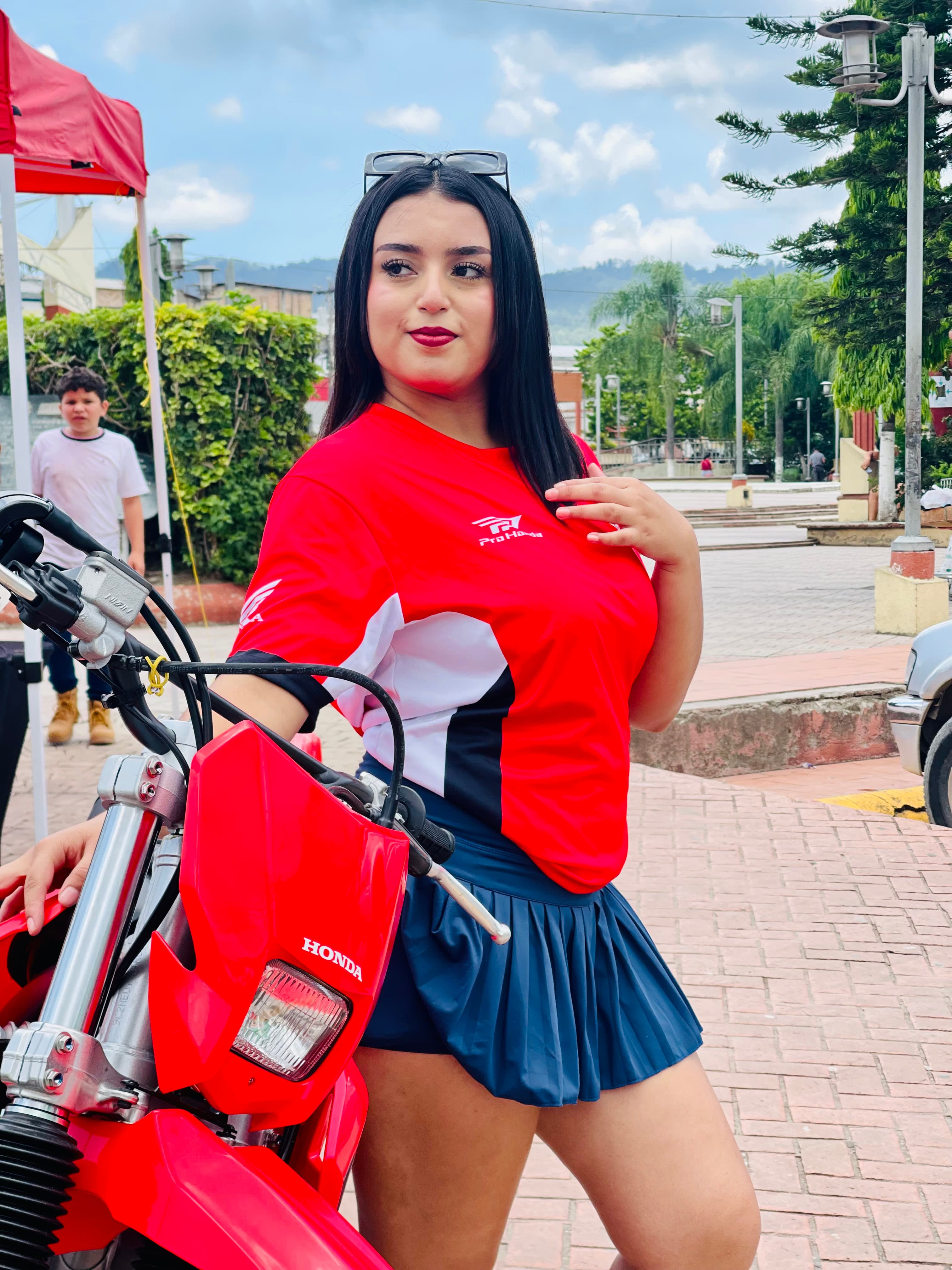 Mujer con camiseta roja Honda y falda azul posando junto a una motocicleta roja.