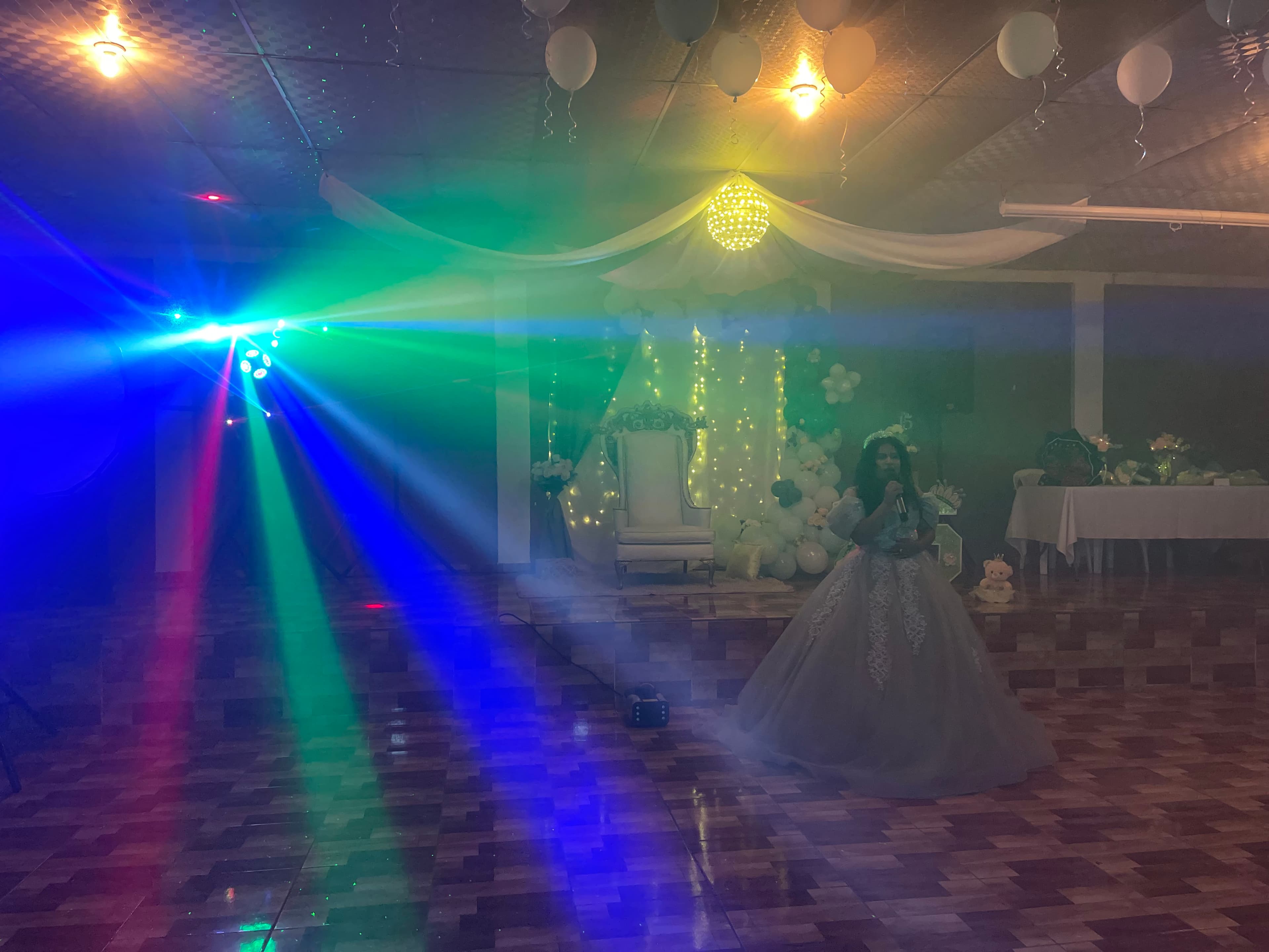 Quinceañera con vestido pomposo canta en un salón iluminado por vibrantes luces de colores.